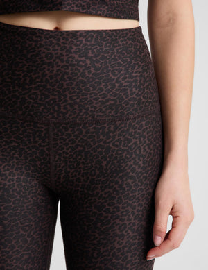 A woman modeling a matching athletic set consisting of a dark brown leopard crop tank and high-waisted leggings, paired with white and gray running shoes, against a light gray studio background.
