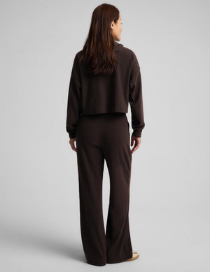 A woman modeling a matching dark brown lounge set consisting of a cropped long-sleeve hoodie with drawstrings and matching wide-leg sweatpants, photographed against a light gray studio background.