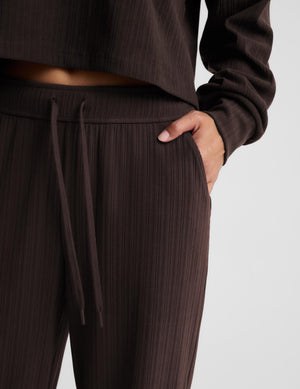 Close-up of a person wearing dark brown ribbed lounge pants with a drawstring waist and a matching cropped long-sleeve top, with one hand in the pocket, against a light gray studio background.