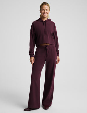 A woman modeling a matching two-piece loungewear set in deep burgundy, featuring a long-sleeve hoodie and wide-leg ribbed pants, against a light gray studio background.