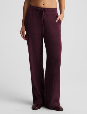 A model wearing a pair of wide-leg, ribbed knit lounge pants in a deep burgundy  paired with dark brown shoes, against a light gray studio background.