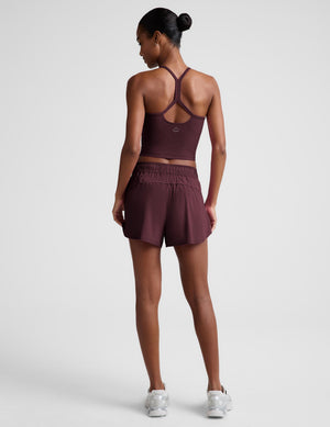 A woman is shown from the back wearing a matching athletic set consisting of a dark plum-burgundy sports bra and shorts, paired with white sneakers, against a light gray studio background.