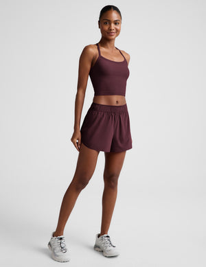 A woman modeling a matching athletic set consisting of a dark plum-burgundy crop top and shorts, paired with white sneakers, against a light gray studio background.