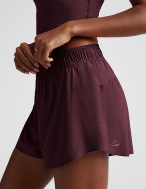 A close-up of a model wearing athletic shorts in a deep plum or burgundy shade. The shorts feature an elastic waistband, a side pocket, and a small white embroidered logo near the hem.