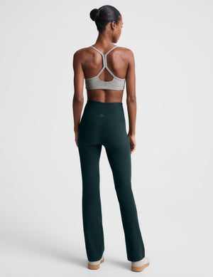 A model wearing dark forest green high-waisted bootcut leggings and a light gray sports bra, standing against a light gray studio background.