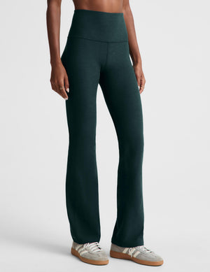 A model wearing dark forest green high-waisted bootcut leggings and a light gray sports bra, standing against a light gray studio background.