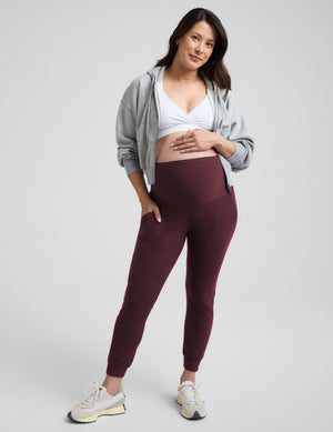 Pregnant woman wearing a grey sweatshirt, white maternity bra, and red maternity joggers against a plain background. 