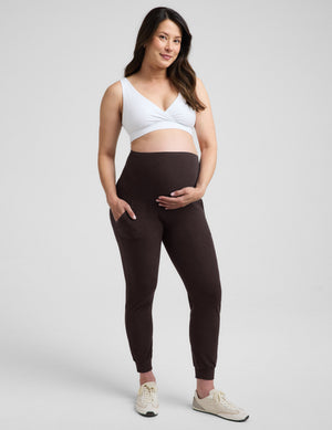 Woman wearing a white nursing bra and brown maternity joggers against a plain background. 