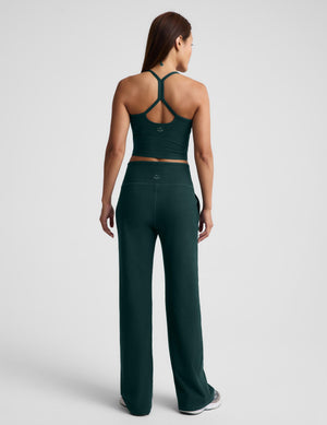 A woman wearing a dark green racerback cropped tank top and dark green wide leg pants against a plain studio background. 