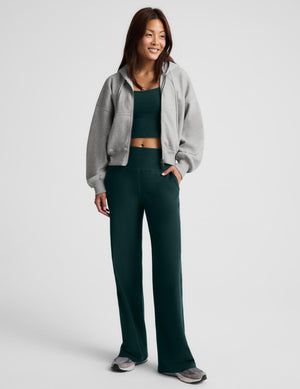 A woman wearing a gray zip-up hooded jacket, dark green racerback cropped tank top and dark green wide leg pants against a plain studio background. 