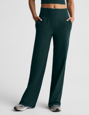 A woman wearing a dark green racerback cropped tank top and dark green wide leg pants against a plain studio background. 