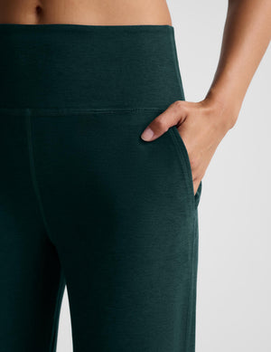 Close-up of a hand in the pocket of dark green wide leg pants against a plain studio background. 