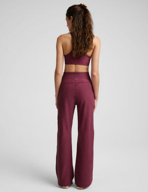 A woman viewed from the back wearing a matching burgundy activewear set consisting of a sports bra and high-waisted wide-leg pants, set against a plain light gray studio background.