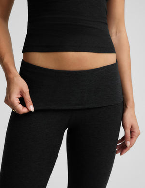 Close-up of a model wearing black high-waisted pants with a foldover waistband, paired with a matching black crop top, against a light gray background.
