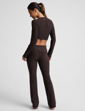 A woman modeling a dark brown two-piece activewear set consisting of a long-sleeve wrap-style crop top and matching high-waisted flared leggings, photographed against a light gray studio background.