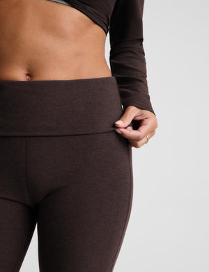 Close-up of a person wearing dark brown high-waisted leggings with a foldover waistband, holding the waistband with one hand. The person is also wearing a matching dark brown long-sleeve top against a light gray studio background.
