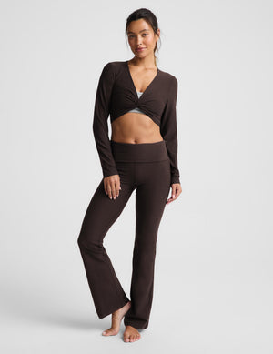 A woman modeling a dark brown two-piece activewear set consisting of a long-sleeve wrap-style crop top and matching high-waisted flared leggings, photographed against a light gray studio background.