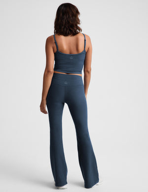 A woman modeling a matching dark blue activewear set consisting of a cropped tank top and high-waisted flared leggings, paired with white sneakers, against a light gray studio background.