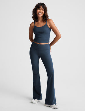A woman modeling a matching dark blue activewear set consisting of a cropped tank top and high-waisted flared leggings, paired with white sneakers, against a light gray studio background.
