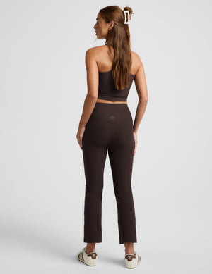 A woman modeling a matching dark brown activewear set consisting of a cropped tank top and high-waisted cropped pants, paired with white sneakers, against a light gray studio background.