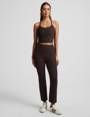 A woman modeling a matching dark brown activewear set consisting of a cropped tank top and high-waisted cropped pants, paired with white sneakers, against a light gray studio background.