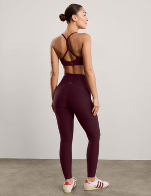 A woman modeling a matching burgundy activewear set consisting of a sports bra and high-waisted leggings, paired with white sneakers, against a neutral studio background.
