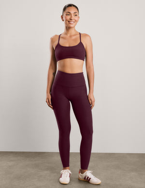 A woman modeling a matching burgundy activewear set consisting of a sports bra and high-waisted leggings, paired with white sneakers, against a neutral studio background.