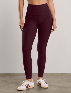 A woman modeling a matching burgundy activewear set consisting of a sports bra and high-waisted leggings, paired with white sneakers, against a neutral studio background.