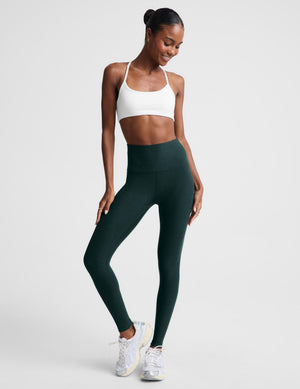 A woman modeling dark forest green high-waisted leggings and a white sports bra against a light gray studio background.
