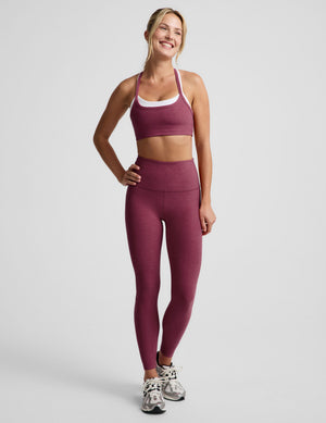 A woman modeling a matching burgundy activewear set consisting of a sports bra with white trim and high-waisted leggings, paired with gray and white sneakers, against a light gray studio background.