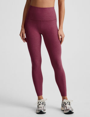 A model wearing high-waisted heathered burgundy leggings and white athletic sneakers, shown from the waist down against a light gray studio background.