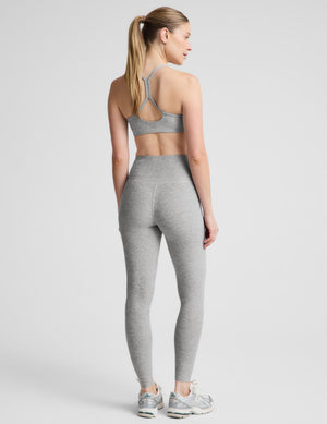A woman modeling a light gray heathered activewear set consisting of a sports bra and high-waisted leggings, paired with gray sneakers, against a solid light gray background.