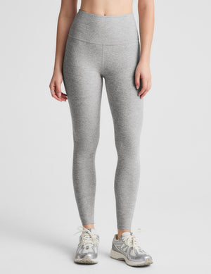 A woman modeling a light gray heathered activewear set consisting of a sports bra and high-waisted leggings, paired with gray sneakers, against a solid light gray background.