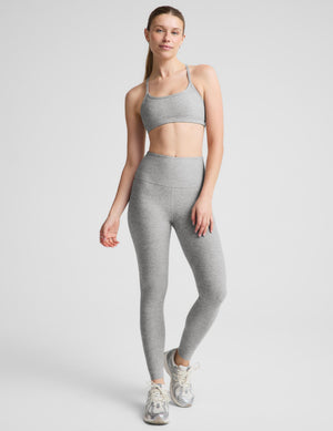 A woman modeling a light gray heathered activewear set consisting of a sports bra and high-waisted leggings, paired with gray sneakers, against a solid light gray background.