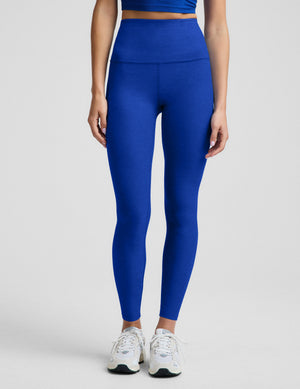A woman modeling a matching royal blue activewear set consisting of a cropped tank and high-waisted leggings, paired with white sneakers, against a light gray studio background.