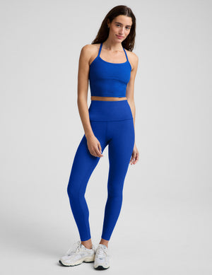 A woman modeling a matching royal blue activewear set consisting of a cropped tank and high-waisted leggings, paired with white sneakers, against a light gray studio background.
