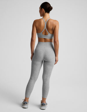 A woman shown from the back wearing a light gray heather sports bra and matching high-waisted leggings, paired with gray and white sneakers, against a light gray studio background.
