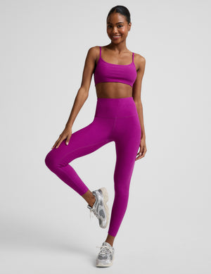 Woman wearing a purple bra and purple high-waisted midi length leggings against a plain background. 