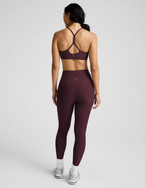 A woman modeling a matching athletic set consisting of a dark burgundy sports bra and high-waisted leggings, paired with white socks and gray sneakers, against a light gray studio background.