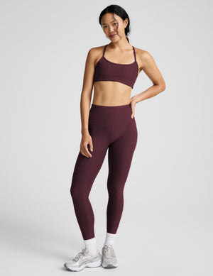 A woman modeling a matching athletic set consisting of a dark burgundy sports bra and high-waisted leggings, paired with white socks and gray sneakers, against a light gray studio background.