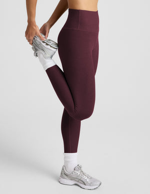 A woman modeling a matching athletic set consisting of a dark burgundy sports bra and high-waisted leggings, paired with white socks and gray sneakers, against a light gray studio background.