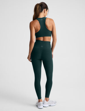 A woman modeling a matching dark green athletic set consisting of a sports bra and high-waisted leggings, paired with white sneakers, against a light gray studio background.