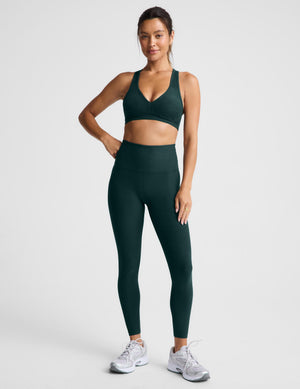 A woman modeling a matching dark green athletic set consisting of a sports bra and high-waisted leggings, paired with white sneakers, against a light gray studio background.