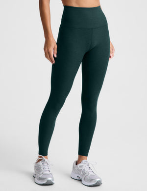 A woman modeling a matching dark green athletic set consisting of a sports bra and high-waisted leggings, paired with white sneakers, against a light gray studio background.