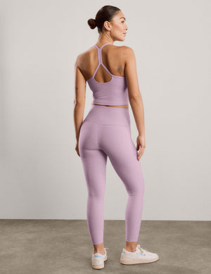 A woman wearing a matching light purple/lavender activewear set, consisting of a sports bra and high-waisted leggings, shown from the back. She is standing in a studio with a light gray wall and concrete floor.