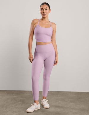 A woman modeling a matching light purple/lavender activewear set consisting of a sports bra and high-waisted leggings, paired with white sneakers, against a neutral studio backdrop.
