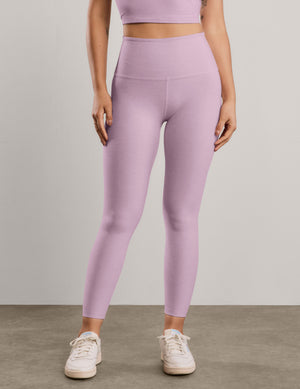 A model wearing high-waisted, ankle-length leggings in a light lavender purple color, paired with a matching sports bra and white sneakers, set against a neutral studio background.