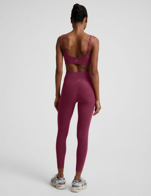 A woman shown from the back wearing a matching burgundy activewear set consisting of a sports bra and high-waisted leggings, paired with gray and white sneakers, against a light gray studio background.