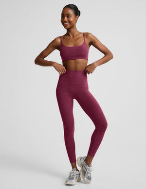 A woman modeling a matching athletic set consisting of a heathered dusty rose sports bra and high-waisted leggings, paired with silver sneakers, against a light gray studio backgroun