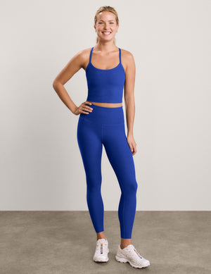 A woman modeling a matching royal blue activewear set consisting of a cropped tank and high-waisted leggings, paired with white sneakers, against a neutral studio backdrop.